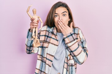 Young caucasian girl holding small wooden manikin covering mouth with hand, shocked and afraid for...