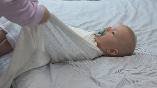 Mother swaddling baby before bedtime, 7 month old infant wrapped on bed, swaddling helps prevent baby from flailing arms and legs. High quality 4k footage