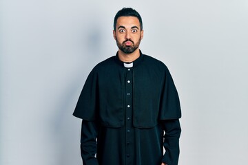 Handsome hispanic man with beard wearing catholic priest robe puffing cheeks with funny face. mouth...