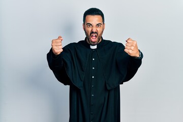 Handsome hispanic man with beard wearing catholic priest robe angry and mad raising fists...