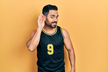 Handsome hispanic man with beard wearing basketball t shirt smiling with hand over ear listening...