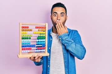 Handsome hispanic man with beard holding traditional abacus covering mouth with hand, shocked and...