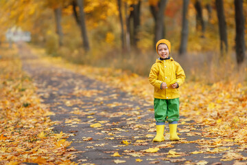 cheerful child boy in yellow raincoat and rubber boots in autumn park
