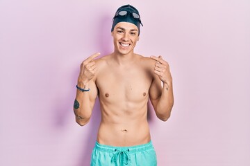 Young hispanic man wearing swimwear and swimmer glasses smiling cheerful showing and pointing with fingers teeth and mouth. dental health concept.