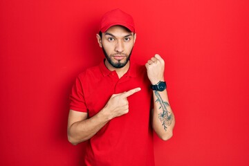 Hispanic man with beard wearing delivery uniform and cap in hurry pointing to watch time,...