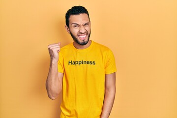Hispanic man with beard wearing t shirt with happiness word message angry and mad raising fist frustrated and furious while shouting with anger. rage and aggressive concept.