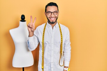 Hispanic man with beard dressmaker designer standing by manikin smiling with happy face winking at...