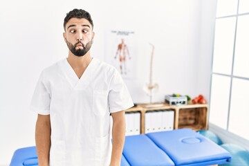 Young handsome man with beard working at pain recovery clinic making fish face with lips, crazy and comical gesture. funny expression.