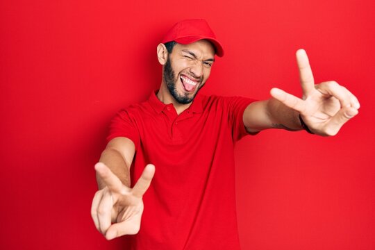 Hispanic man with beard wearing delivery uniform and cap smiling with tongue out showing fingers of both hands doing victory sign. number two.