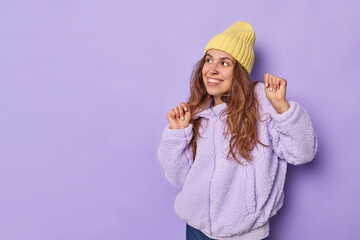 Horizontal shot of happy long haired woman keeps hands raised up smiles gently dances and feels pleased dressed in artificial fur coat and hat poses against purple background with blank space