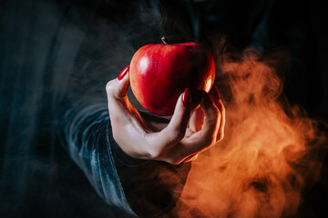 Woman as witch in black offers red apple as symbol of temptation, poison. Fairy tale, white snow wizard concept. Spooky halloween, cosplay. Smoke, haze background. - obrazy, fototapety, plakaty