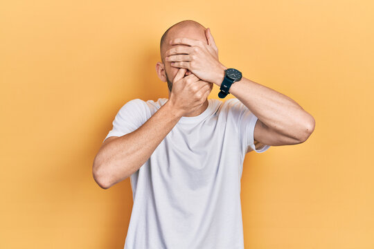 Young bald man wearing casual white t shirt covering eyes and mouth with hands, surprised and shocked. hiding emotion