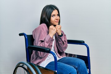 Young brunette woman sitting on wheelchair ready to fight with fist defense gesture, angry and upset face, afraid of problem