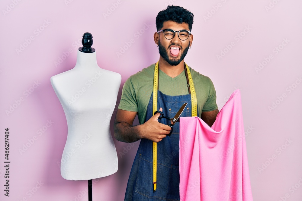 Poster arab man with beard dressmaker designer holding scissors and cloth sticking tongue out happy with fu