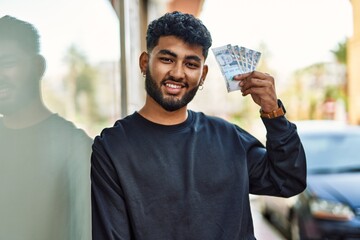 Young arab man smiling confident holding sol peruvian banknotes at street - obrazy, fototapety, plakaty