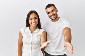 Obraz premium Young interracial couple standing together in love over isolated background smiling friendly offering handshake as greeting and welcoming. successful business.