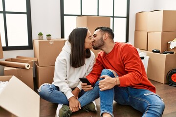 Young latin couple kissing and using smartphone at new home.