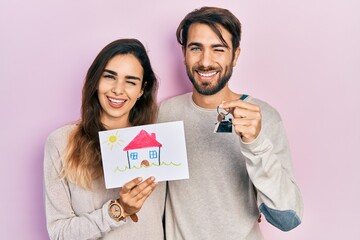Young hispanic couple holding house draw and key of new home winking looking at the camera with...