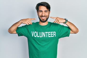 Young hispanic man wearing volunteer t shirt smiling cheerful showing and pointing with fingers teeth and mouth. dental health concept.