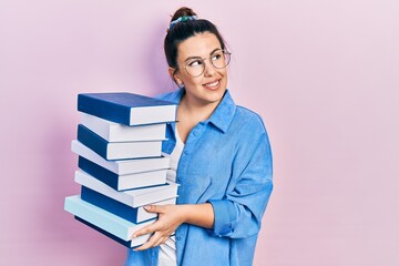 Young hispanic woman wearing glasses and holding books smiling looking to the side and staring away thinking.