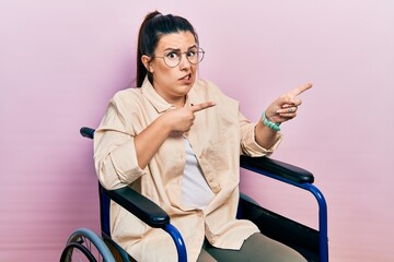 Young hispanic woman sitting on wheelchair pointing aside worried and nervous with both hands,...