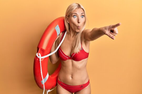 Young blonde woman wearing bikini and holding lifeguard float pointing with finger afraid and shocked with surprise and amazed expression, fear and excited face.