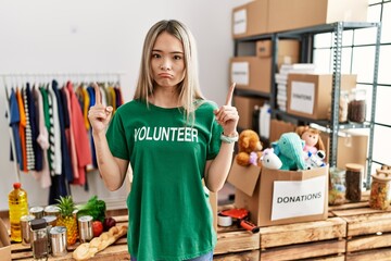 Asian young woman wearing volunteer t shirt at donations stand pointing up looking sad and upset,...