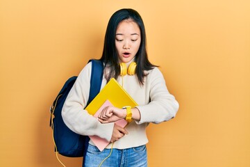 Young chinese girl holding student backpack and books looking at the watch time worried, afraid of...
