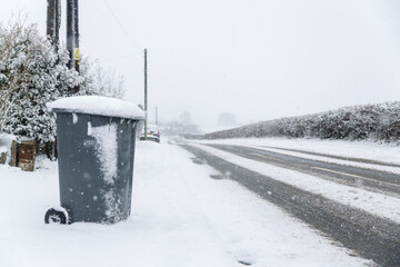 A council wheelie bin sitting by the side of the road in a snow covered landscape waiting for the weekly collection