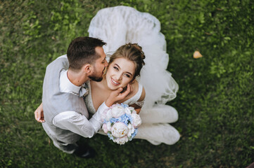 Stylish young, bearded groom in a gray suit and a beautiful, sweet bride in a white dress hug and dance against the background of green grass in nature. Wedding portrait of happy newlyweds.