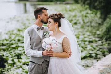 Stylish, fashionable, bearded groom in a gray suit and bow tie hugs and kisses a beautiful, smiling bride in a white dress on the background of a lake with green lily leaves. Wedding photography.