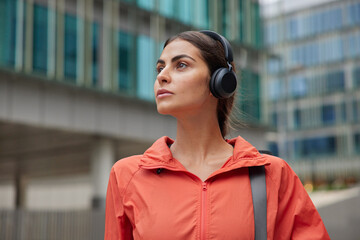 Photo of young active brunette woman strolls outdoors listens music via headphones wears windbreaker carries karemat poses against outdoor against blurred city background. leads healthy lifestyle © WHstudio Leushin N