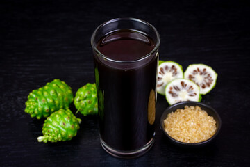 Noni juice in transparent glass and fresh noni fruit with sugar cane on small black bowl on black...