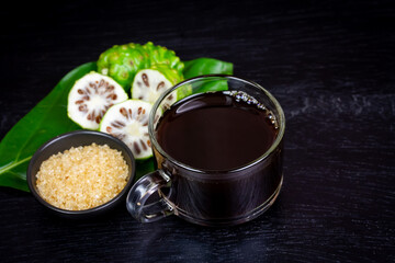 Noni juice in transparent glass cup and fresh noni fruit with green leaf on black wooden background.