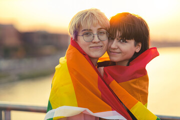 Young non binary couple in love hugging under a rainbow flag 
