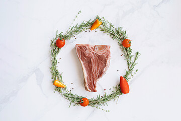 raw t-bone steak in the center of a Christmas wreath of rosemary thyme cherry tomatoes and bell peppers on a white marble surface. christmas dinner concept