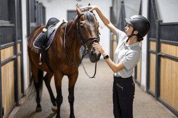 Female horseman with her brown Thoroughbred horse in stable. Concept of animal care. Rural rest and...