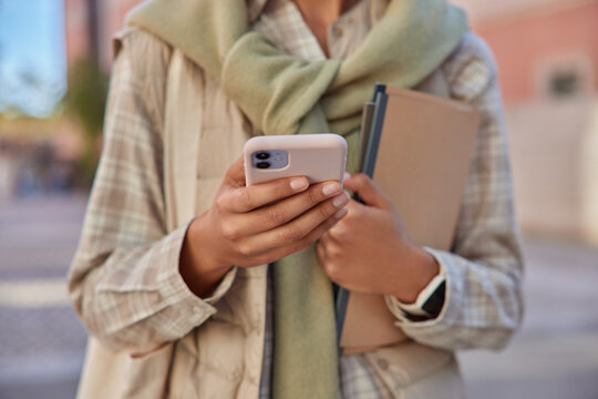 Cropped image of unknown woman in casual clothes holds mobile phone browses web publication in social media holds paper notepad poses outdoors against blurred background messaging and walking in city