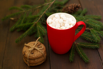 On a dark wooden background, a red cup with cocoa and marshmallows and cookies, decorated with fir branches