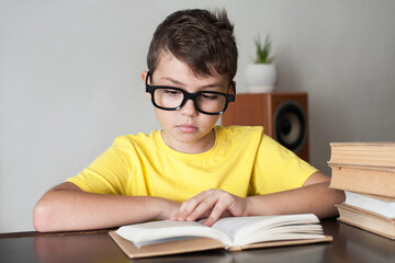 a boy with glasses and a yellow T-shirt reads books at the table at home. Home education