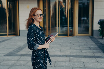 Skilled redhead businesswoman stands sideways has cheerful expression carries modern gadgets