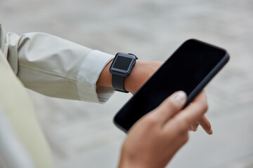 Unrecognizable woman uses modern devices for sport. Cropped shot of womans hand with wearable smartwatch and mobile phone empty screen checking fitness results and monitoring physical activity