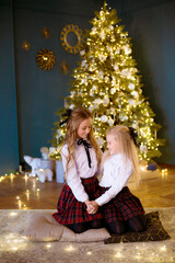 Lovely girls sisters with blonde curly hair with bans to the tols in classic school clothes by the Christmas tree  with presents in a classic interior. Christmas mood 