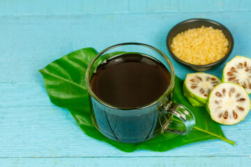 Noni juice in transparent glass cup and fresh noni fruit with green leaf on blue wooden background.