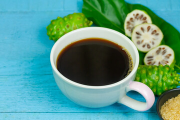 Noni juice in ceramic cup and fresh noni fruit with green leaf on blue wooden background.