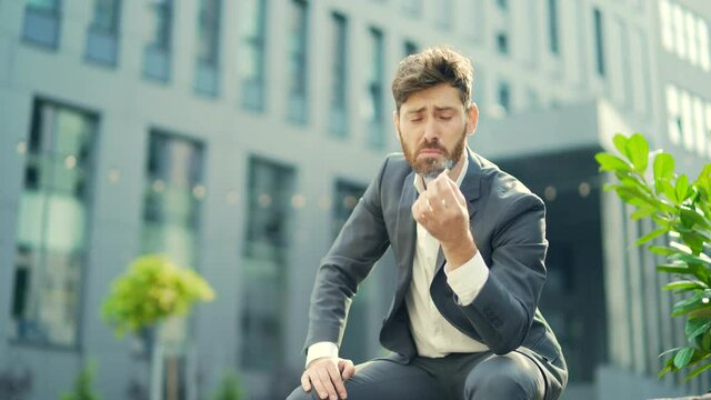 Caucasian bearded business man smoking cannabis outdoors sitting on city park bench on urban street background. Male employee Businessman. Office worker in suit Relieves stress a marijuana outside