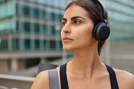 Horizontal shot of thoughtful sporty curly woman looks away has pensive expression walks outdoors carries fitness mat prepares for workout poses outdoors against blurred background listens music
