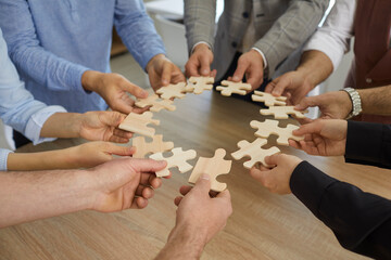 Team of business people standing around office table joining pieces of jigsaw puzzle, close up, closeup shot. Group of entrepreneurs cooperating and forming coalition or alliance. Teamwork concept