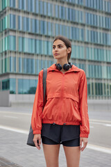 Vertical shot of thoughtful sporty woman dressed in activewear carries fitness mat going to have outdoor training poses on road against modern city building. Athletic female trainer strolls in city © WHstudio Leushin N