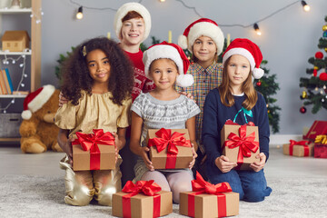 Group of happy smiling multiracial children looking at camera and holding presents tied with red bows, sitting in cozy decorated living-room on Christmas morning at home or during party with friends